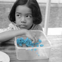 A little girl eaeting popcorn served on a wooden dining table; frowning, glancing, unhappy, upset.