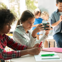 Diverse multiethnic kids students using smartphones in classroom. Multicultural children holding devices having fun with mobile phones apps playing games and checking social media at school.