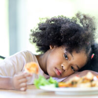 A young, curly haired African American girl sits bored looking at food. Do not want to eat food.
