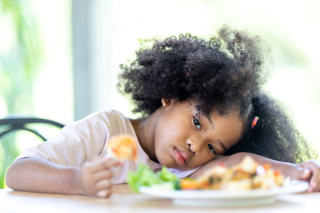 A young, curly haired African American girl sits bored looking at food. Do not want to eat food. 
