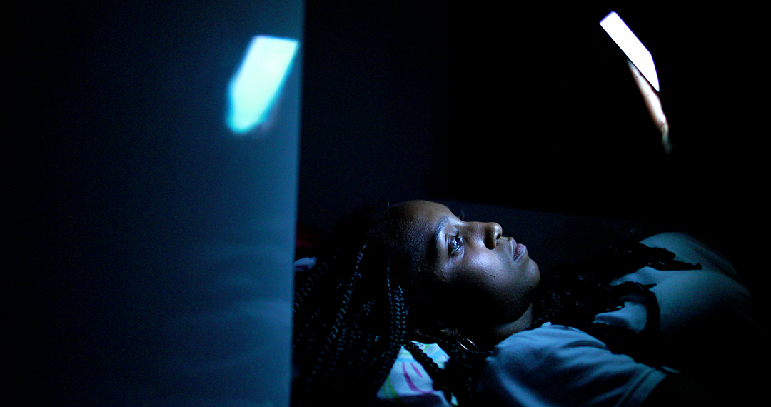 Black girl looking at cellphone screen at night lying in bed with bright blue light