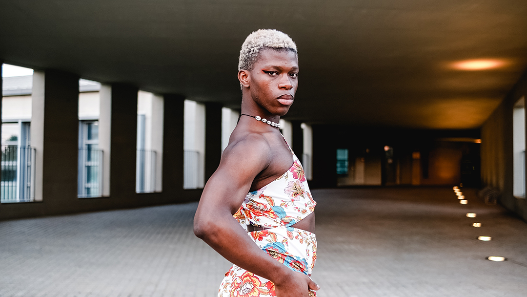 Side view of self assured young African American androgynous male with short dyed hair and makeup in stylish dress standing on street and looking at camera with confidence