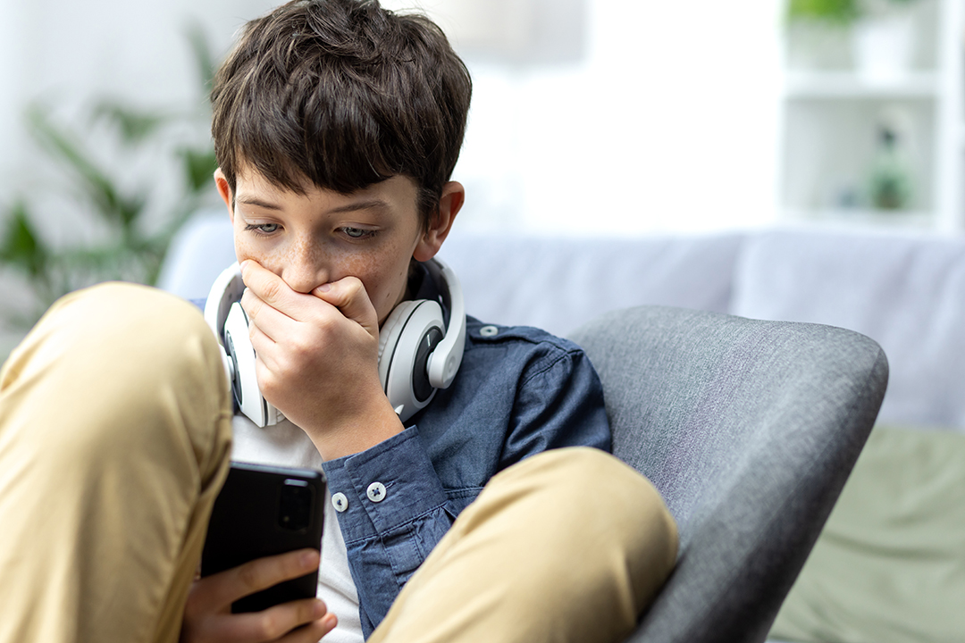 Young teenager boy reading bad news on phone, son sitting on sofa in living room with headphones at home closeup.