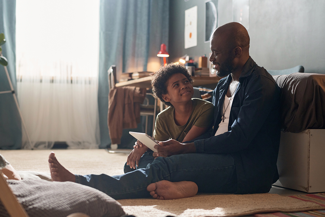 Full length portrait of African American single father and smiling son using tablet and chatting sitting on floor in harsh sunlight copy space