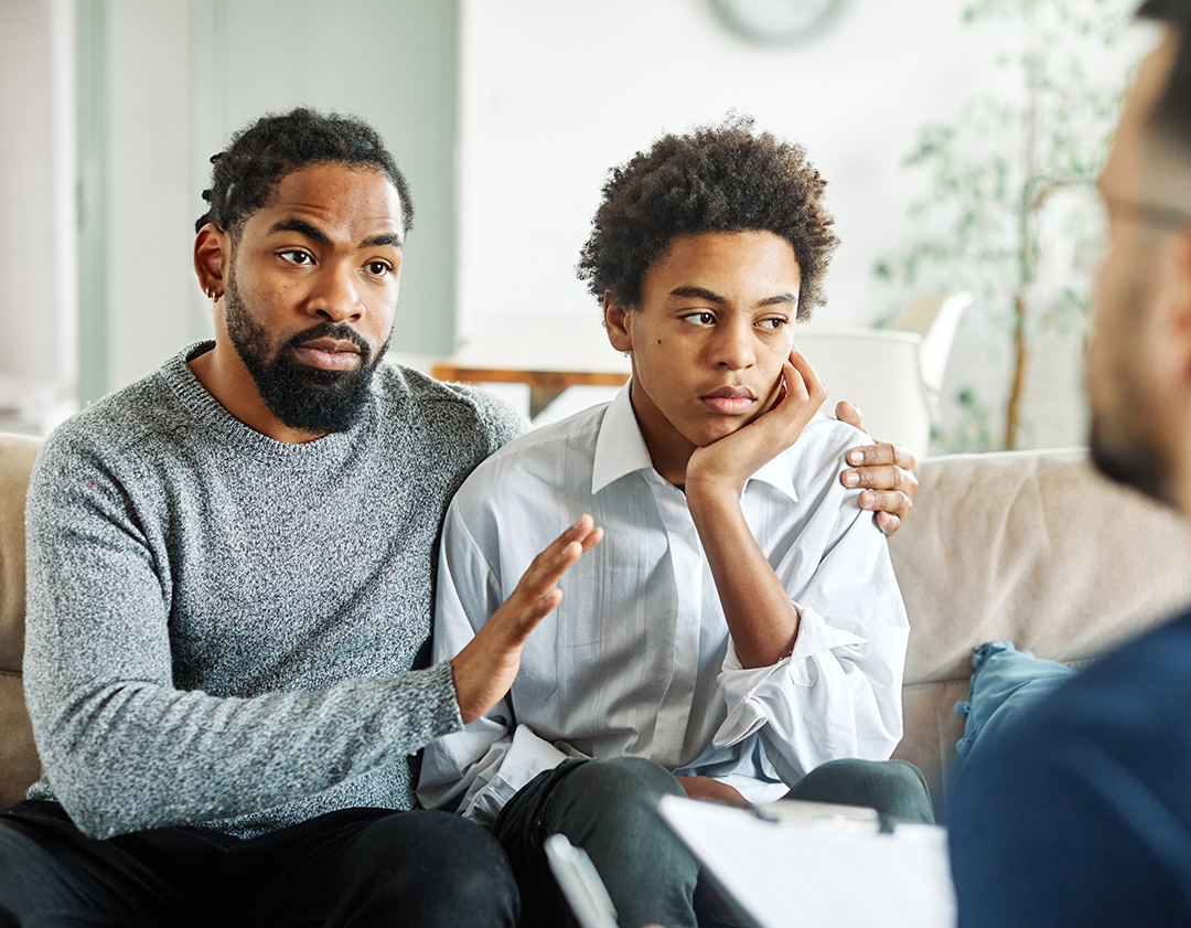 Father with his teenage son at meeting with social worker, psychologist discussing mental health family sitting on sofa in psychotherapist office