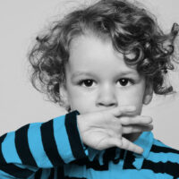 Beautiful curly toddler looking forward and holding his hand on his mouth, boy trying not to speak, shut up