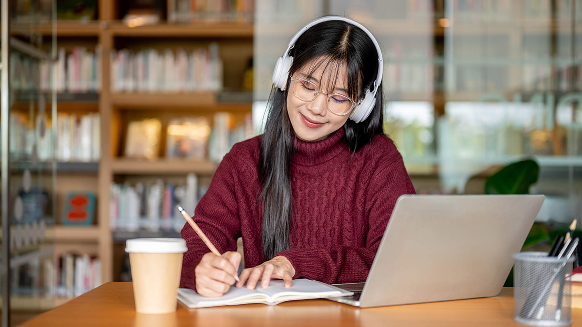 girl sat at desk wearing headphones writing on a notebook with a laptop in front of her
