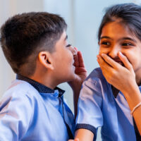 Indian School Kids Sharing Gossip with Friend in a Classroom While Wearing School Uniforms, Whispering Secret Conversations and Laughing Quietly During Leisure Moment Between Study Sessions in school