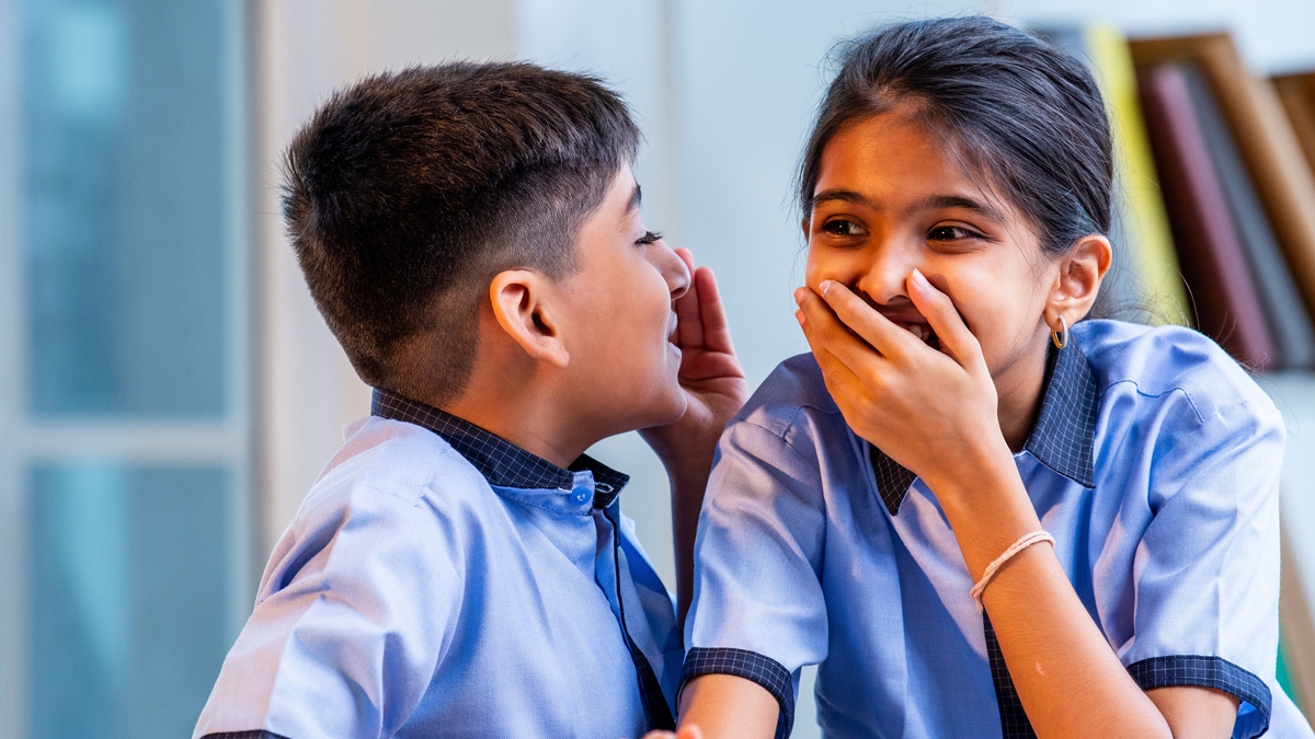 Indian School Kids Sharing Gossip with Friend in a Classroom While Wearing School Uniforms, Whispering Secret Conversations and Laughing Quietly During Leisure Moment Between Study Sessions in school