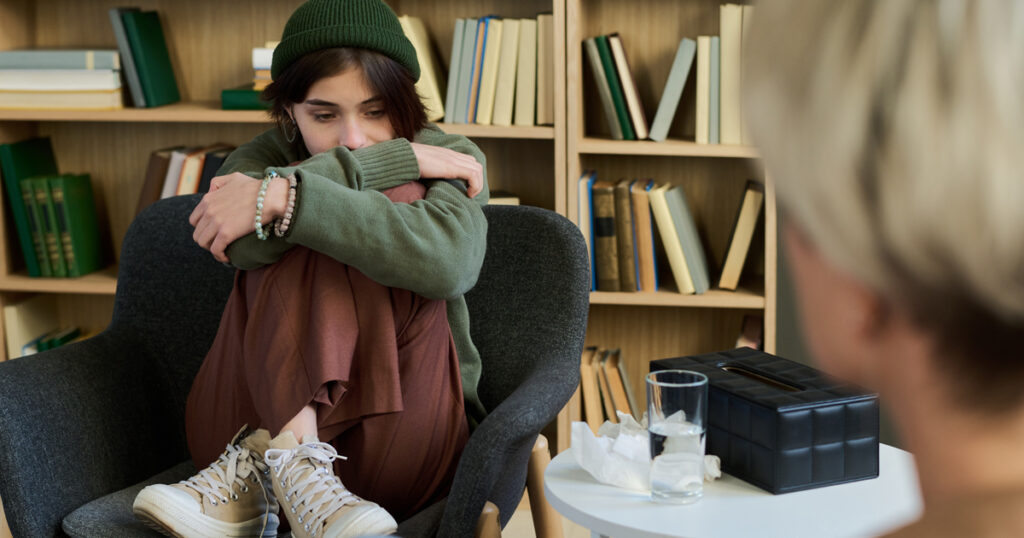 Teenage girl sitting curled up in chair hugging knees during group therapy session, looking down with thoughtful expression while adult woman listening nearby