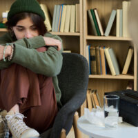 Teenage girl sitting curled up in chair hugging knees during group therapy session, looking down with thoughtful expression while adult woman listening nearby
