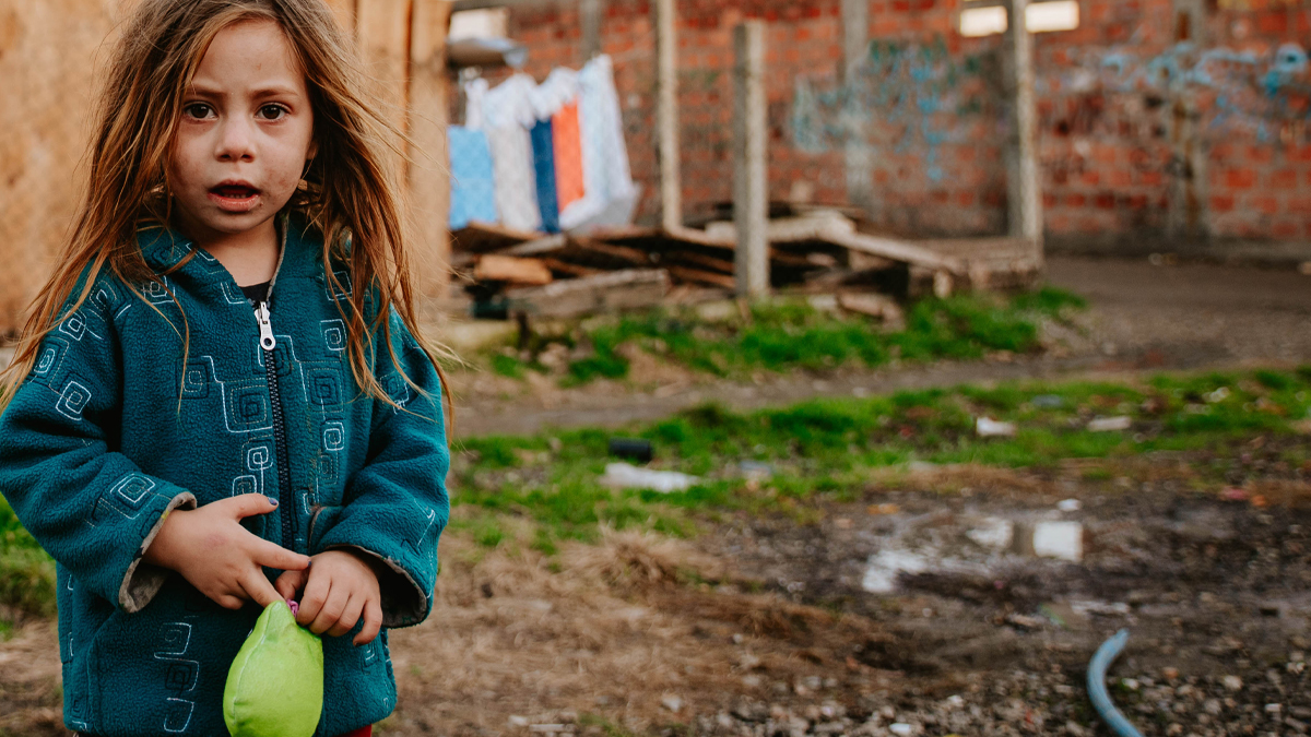 A poor and sad little girl is standing in a Roma settlement, holding a toy in her hand and looking at the camera A poor and sad little girl is standing in a Roma settlement, holding a toy in her hand and looking at the camera