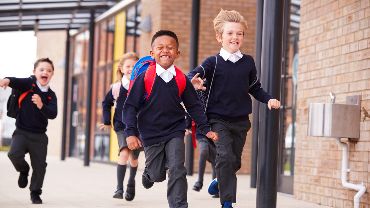 Happy primary school kids, wearing school uniforms and backpacks, running on a walkway outside their school building, front view, close up Happy primary school kids, wearing school uniforms and backpacks, running on a walkway outside their school building, front view, close up
