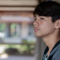 Pensive side portrait of a teen boy in a grey hoodie, leaning against a pillar in an outdoor walkway
