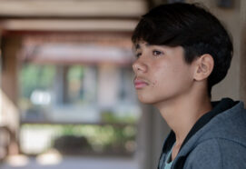 Pensive side portrait of a teen boy in a grey hoodie, leaning against a pillar in an outdoor walkway