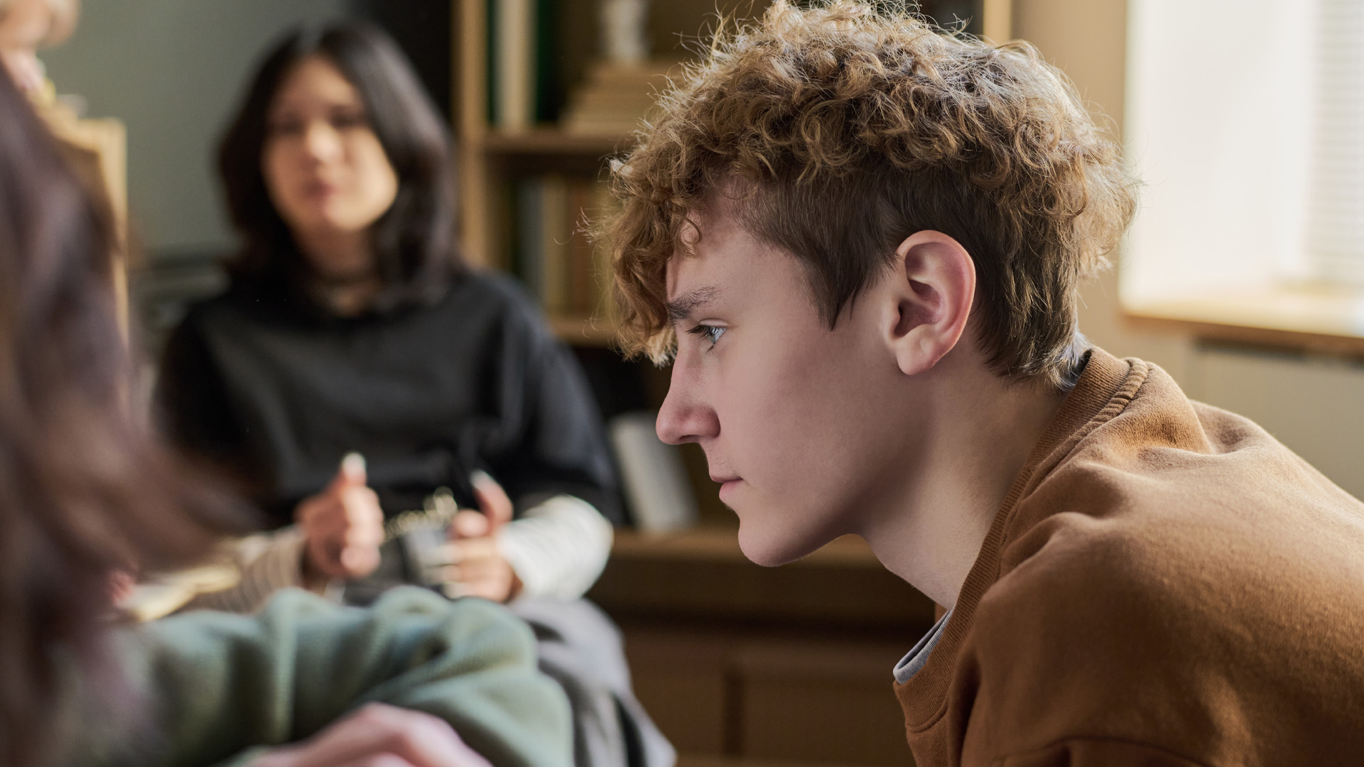 Teenager boy sitting in profile listening attentively within a group with multiethnic young people and teenagers in background participating in discussion