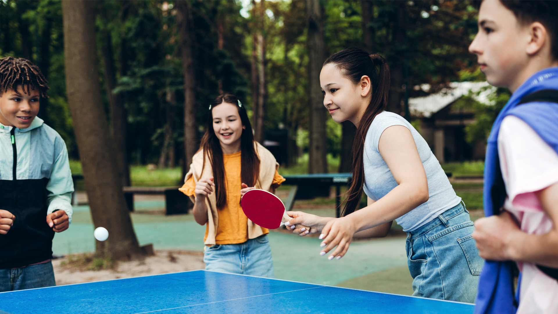 Group of happy teenagers playing table tennis outdoors in a green park. 