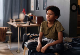 Full length portrait of teenage boy sitting on bed in his room and looking away