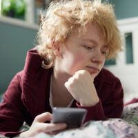 Unhappy Teenage Girl Lying On Bed At Home With Mobile Phone